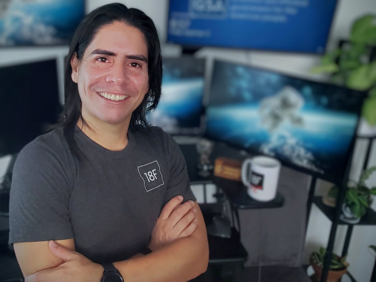 Smiling man with long dark hair, wearing a gray '18F' t-shirt, standing with arms crossed in a tech-filled workspace with multiple monitors displaying space-themed images.