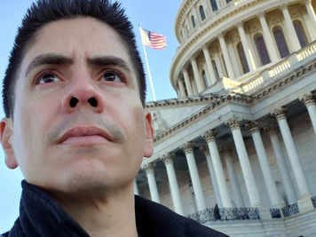 Close-up of a man in a black jacket with a serious expression, standing in front of the U.S. Capitol, with the American flag waving behind him.