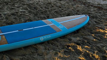A turquoise BOGA stand-up paddleboard resting diagonally on warm beach sand at golden hour.