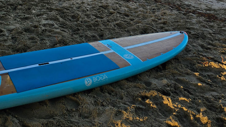 A turquoise BOGA stand-up paddleboard resting diagonally on warm beach sand at golden hour.