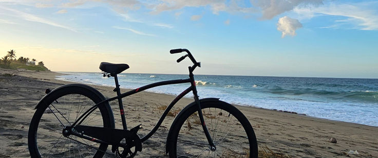 A black beach cruiser bicycle parked on the sand at golden hour, ocean and soft cloud horizon behind it.