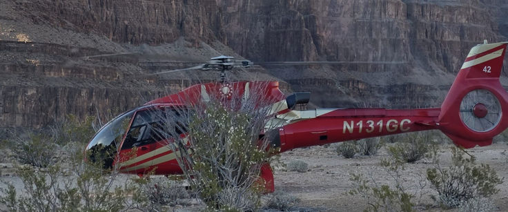 A red Eurocopter EC130 helicopter (N131GC) parked on the desert floor of the Grand Canyon, towering red-rock cliffs rising in the background at dusk.