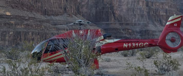 A red Eurocopter EC130 helicopter (N131GC) parked on the desert floor of the Grand Canyon, towering red-rock cliffs rising in the background at dusk.